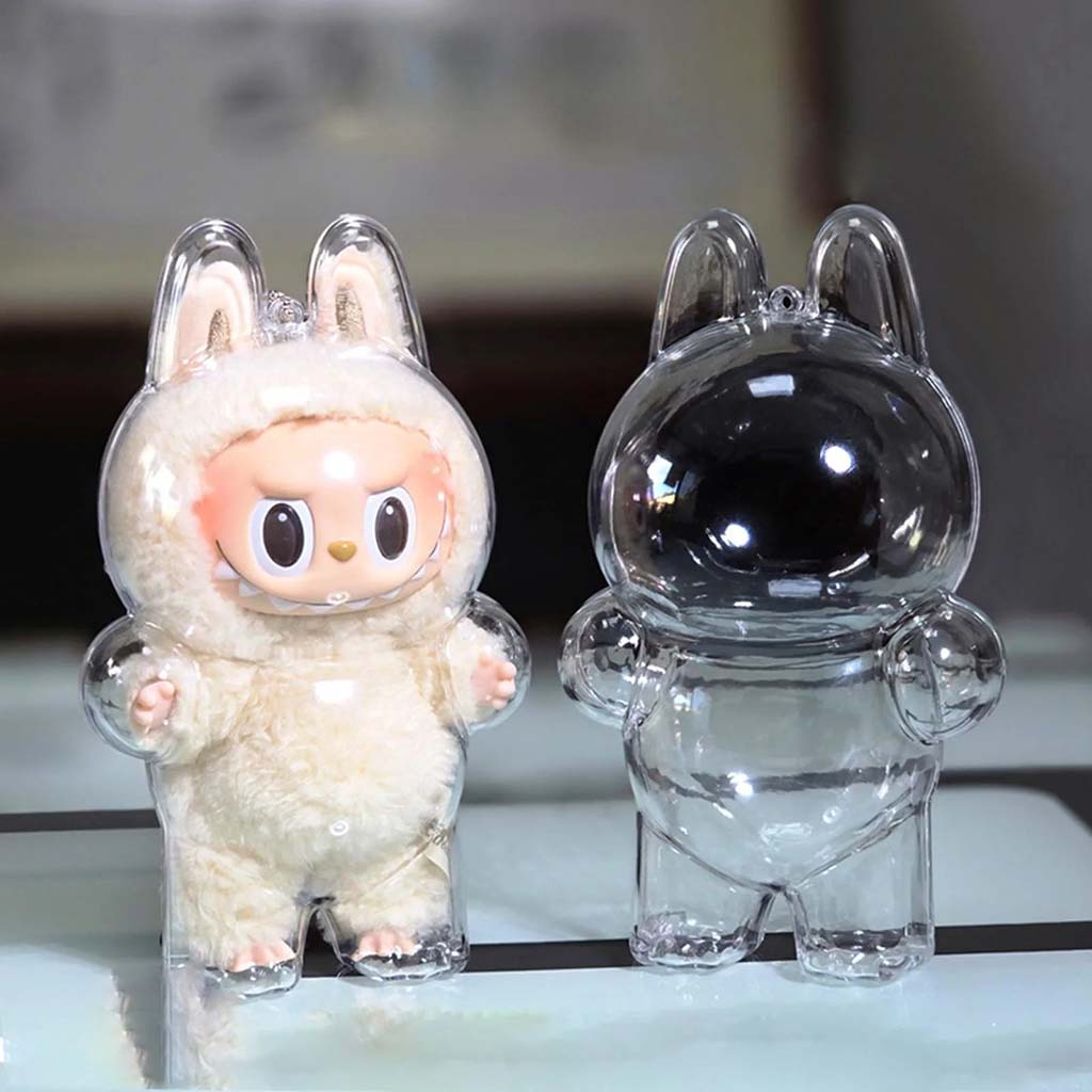 Labubu plush figure in a white fluffy costume beside an empty clear plastic display case shaped like its body, both placed on a reflective surface.