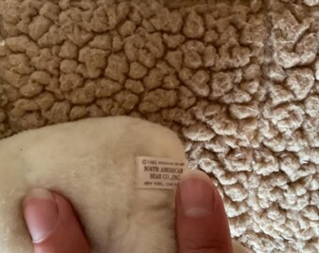 Close-up of a plush toy tag reading “North American Bear Co., Inc.” held by fingers against a textured fabric background.