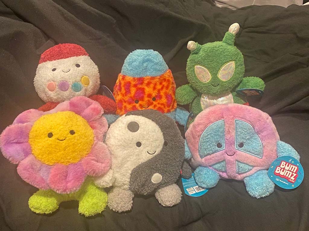 A group of six soft BumBumz plush toys in various fun designs including a flower, alien, peace sign, rocket, rainbow, and yin-yang character, displayed on a dark background.