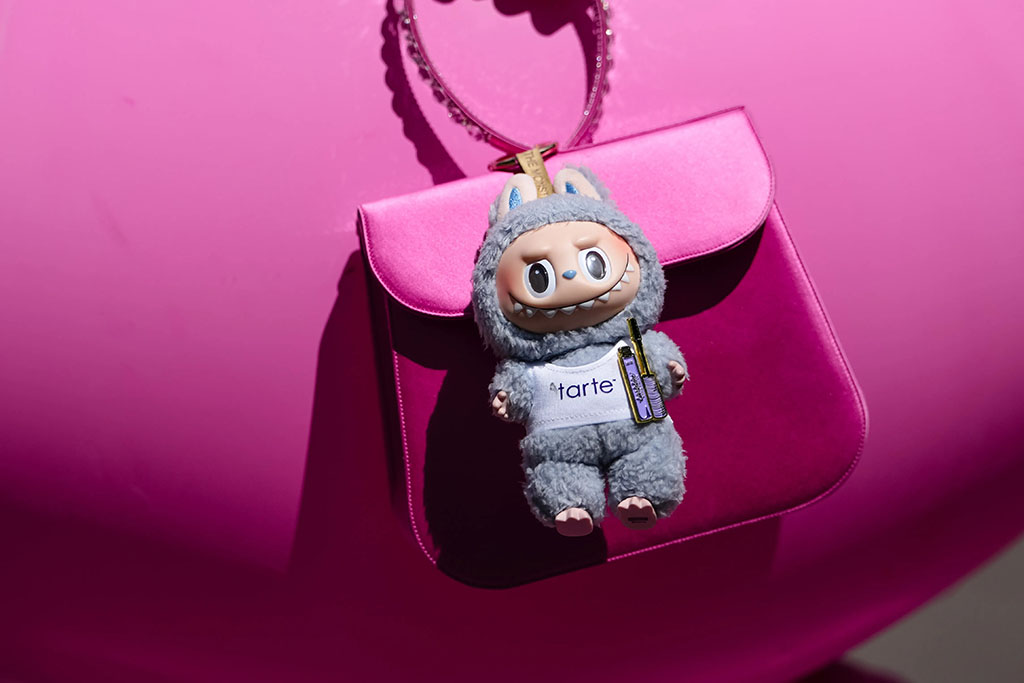 Gray Labubu plush doll wearing a white “tarte” outfit hanging on a shiny pink handbag against a bright pink background.