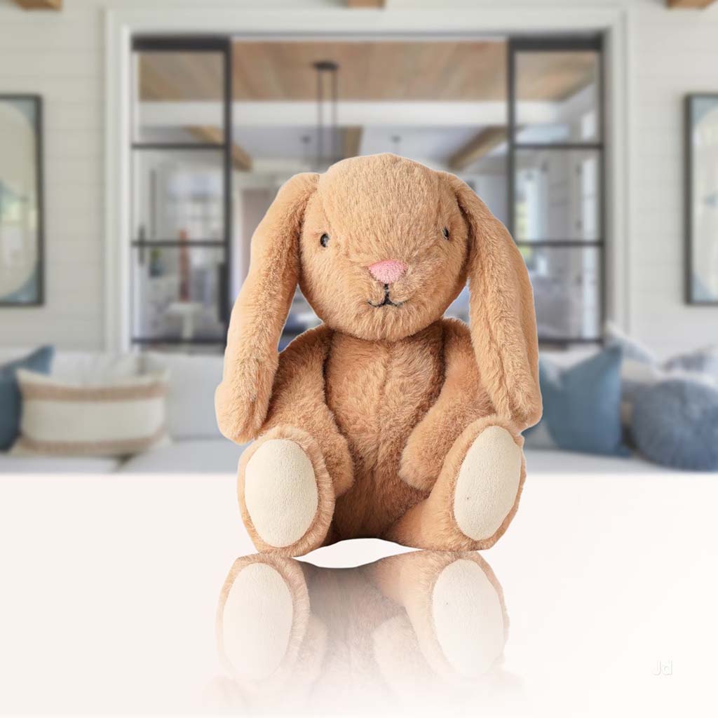 Brown plush bunny toy with long floppy ears sitting upright on a reflective surface, with a cozy modern living room blurred in the background.