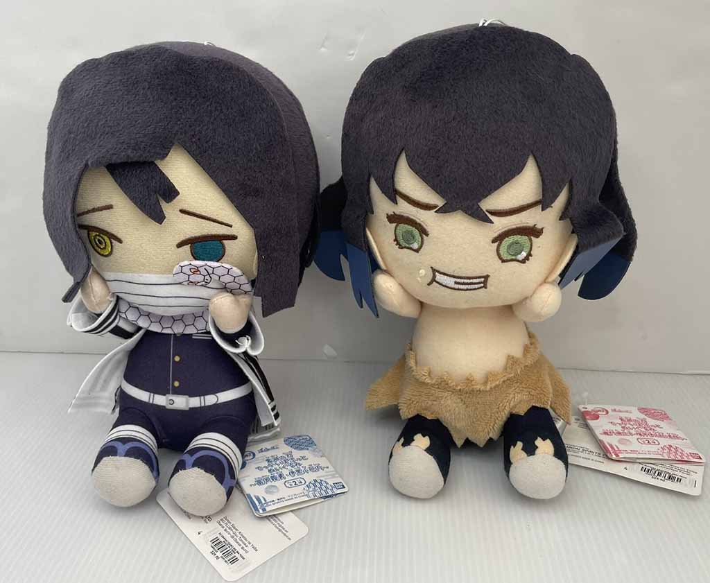  Two cute anime-style plush dolls sitting side by side, one dressed in dark clothing with a white scarf and the other wearing a tan outfit with blue hair and green eyes.