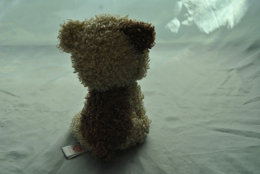 Soft brown and cream curly-textured plush toy sitting with its back facing the camera on a light fabric surface, showing its fluffy material and stitched tag.