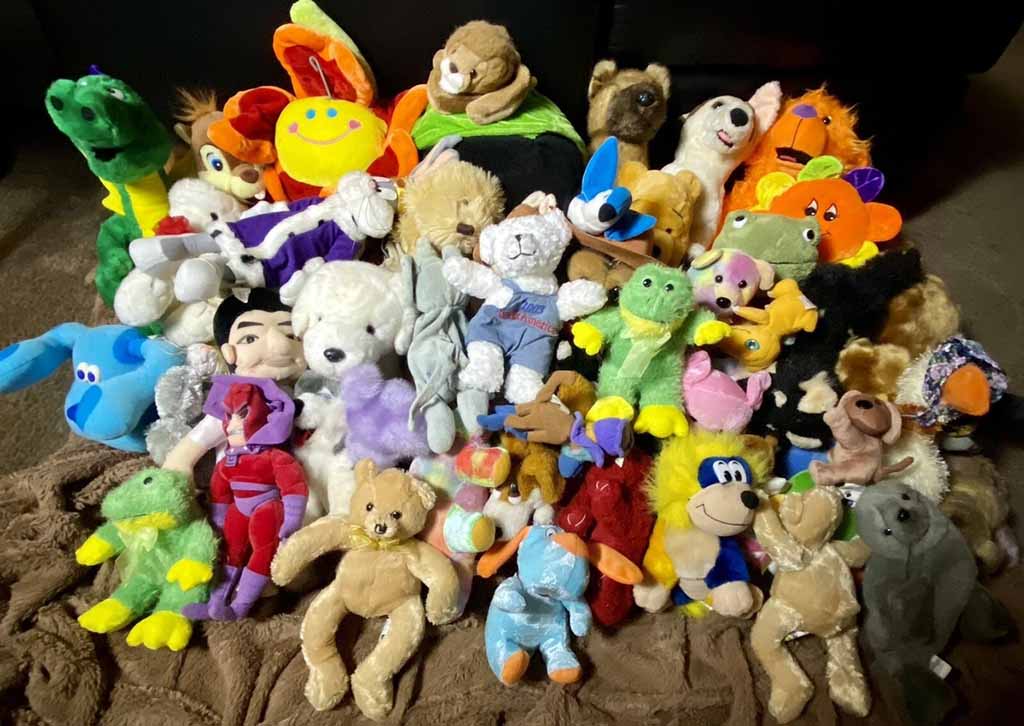 A large assorted collection of plush toys arranged closely together on a blanket, featuring animals, characters, colorful creatures, and teddy bears in various sizes, shapes, and bright colors.