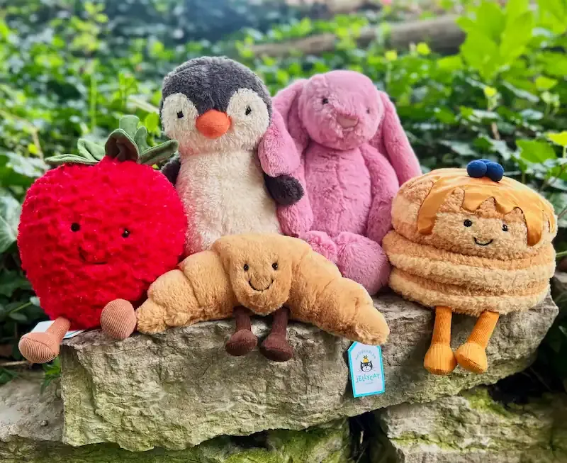 A group of Jellycat plush toys—including a red fruit, penguin, pink bunny, croissant, and pancake—sitting on a stone ledge outdoors with greenery in the background.