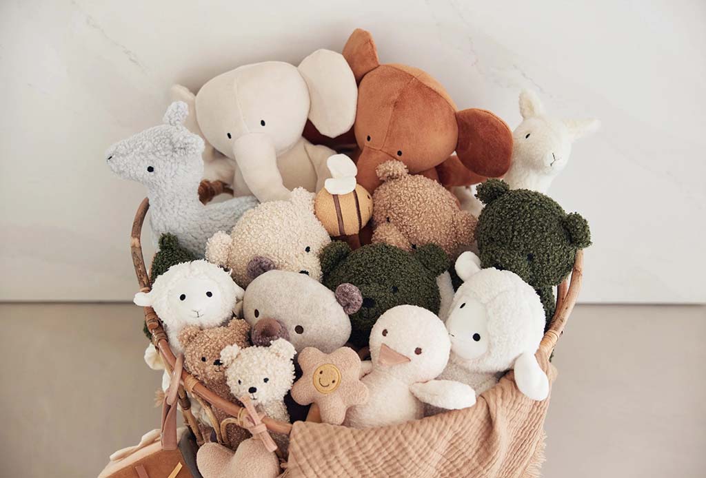 A woven basket filled with soft plush toys including bears, elephants, lambs, and other cuddly animals in neutral and earthy colors, arranged in a cozy home setting.