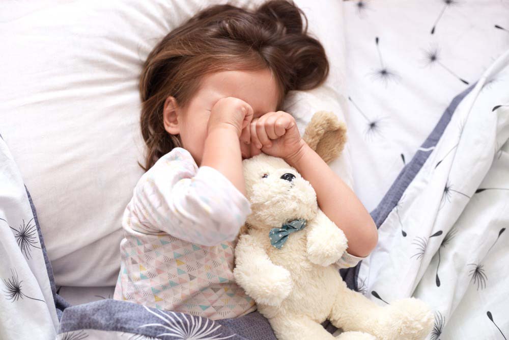 Young child rubbing eyes while lying in bed and holding a soft white plush toy with a blue bow.