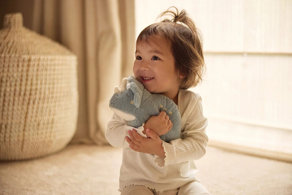 A smiling toddler sitting on the floor and hugging a small blue dinosaur plush toy, showing the warmth and comfort soft toys bring to young children.