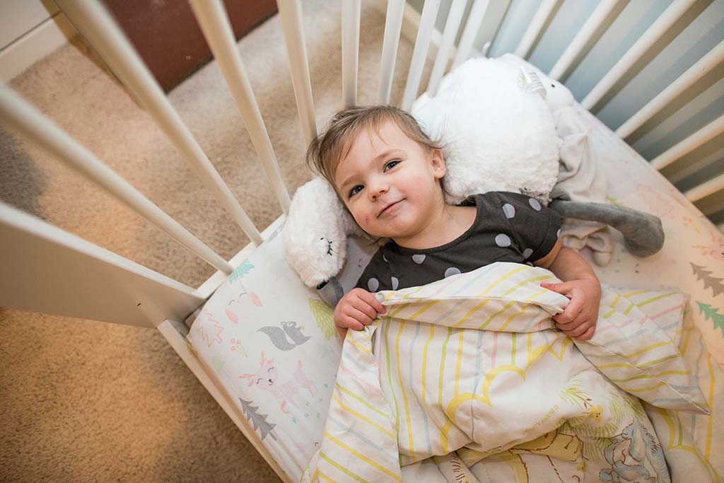 A smiling toddler lying in a crib, covered with a striped blanket and cuddling a soft plush toy, surrounded by light-colored bedding with animal patterns.