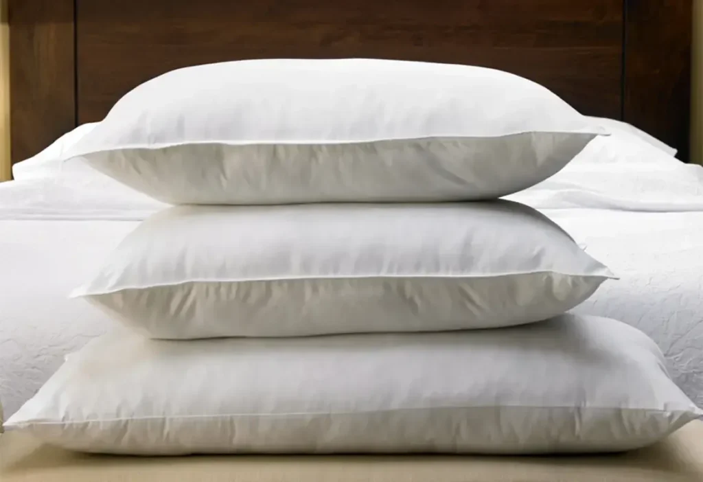 Three white down-alternative bed pillows stacked neatly on a bed with a wooden headboard in a minimal, clean bedroom setting.