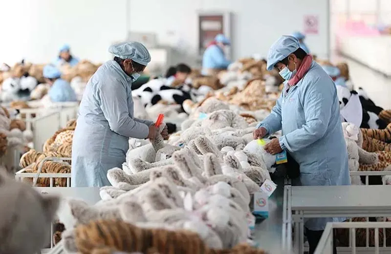 Workers in protective uniforms inspecting and grooming plush toys inside a large stuffed-animal manufacturing factory.
