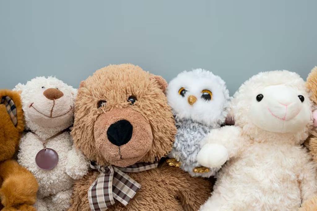 A close-up lineup of various plush animals including a teddy bear, owl, lamb, and other soft toys arranged side by side against a light grey background.