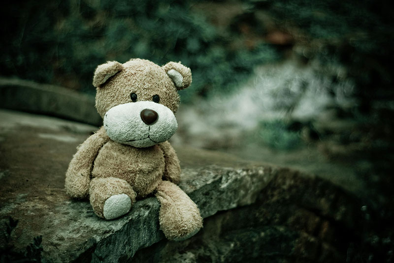 A worn brown teddy bear sits alone on a stone ledge outdoors, surrounded by a dark, moody, blurred forest background.