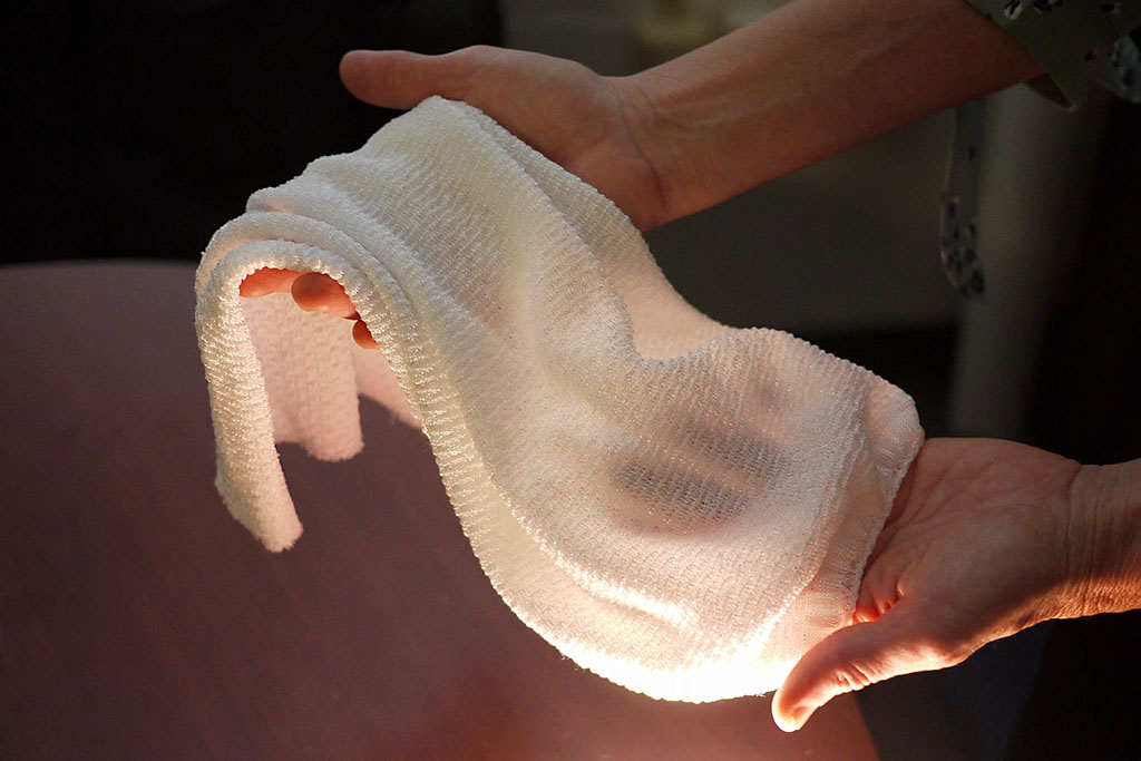 Hands holding a flexible textured white fabric sample, showing its softness and drape under light.