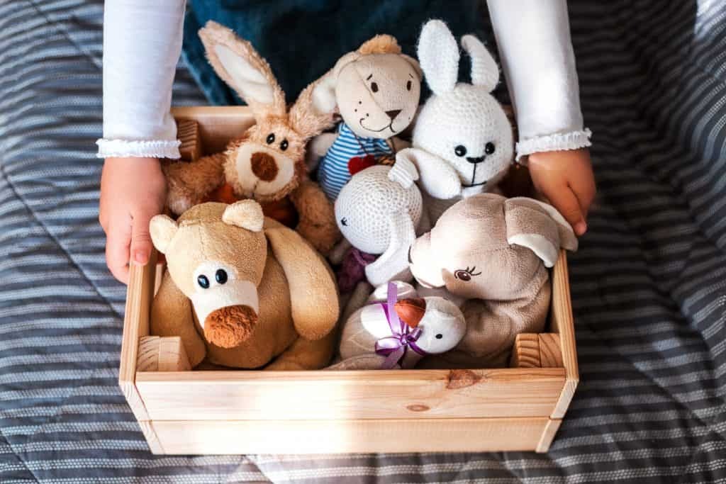Child holding a wooden box filled with assorted stuffed animals, including bears, bunnies, and crocheted plush toys.