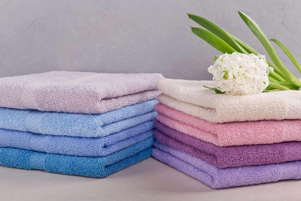 Neatly folded pastel terry cloth towels in blue, pink, and purple shades with a white flower on top, showing soft and absorbent fabric texture.
