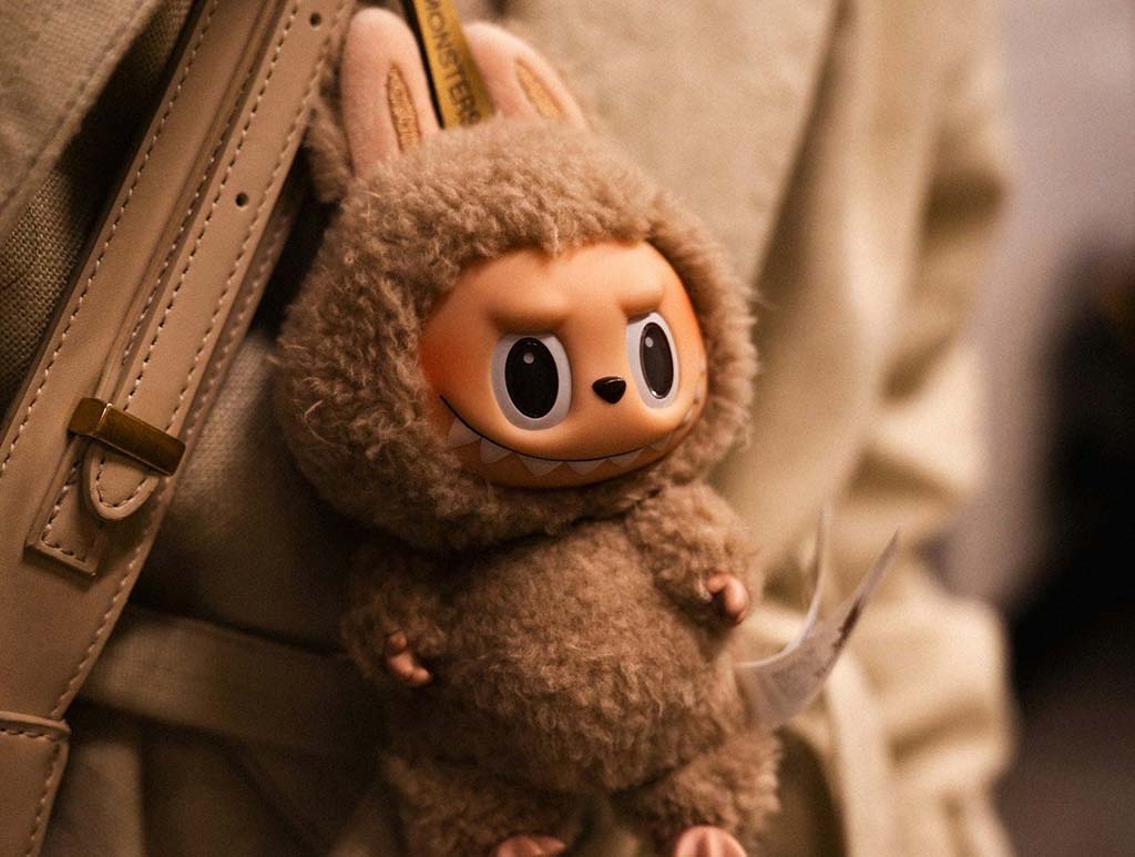 Close-up of a collectible designer plush doll with a fuzzy costume and expressive plastic face clipped to a beige bag.