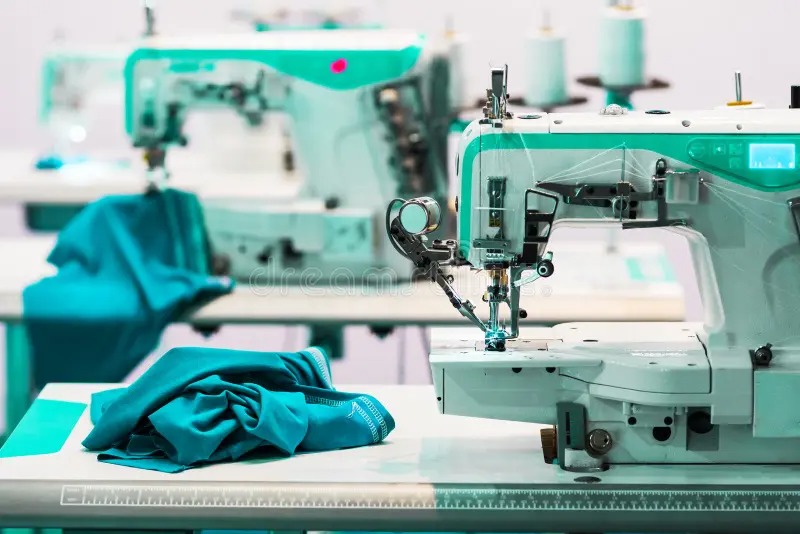 Close-up of industrial sewing machines in a textile factory with teal fabric placed on the worktable, showing professional equipment used for stitching plush toys and garments.