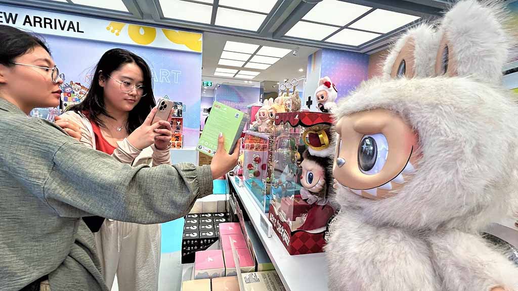 Two women examine and photograph collectible toy boxes in a store, standing beside a large white Labubu figure and shelves filled with POP MART figurines and plush characters.