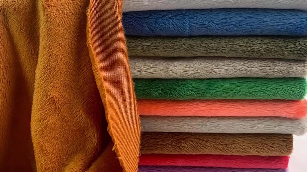 A stack of soft plush fabrics in multiple colors, including orange, blue, green, pink, gray, and brown, showing texture and thickness for plush toy manufacturing.