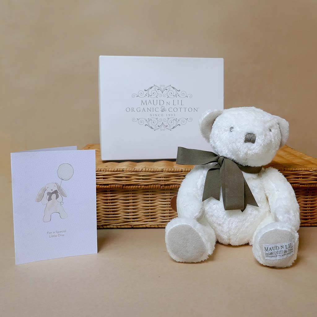 A soft white organic-cotton teddy bear with a dark ribbon sits beside a woven gift basket, a white Maud N Lil gift box, and a small illustrated greeting card on a neutral background.