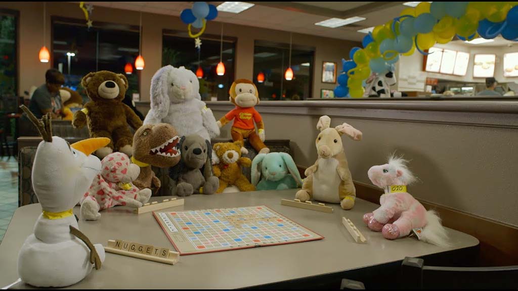Group of plush toys arranged around a restaurant table playing Scrabble under festive blue and yellow balloons.