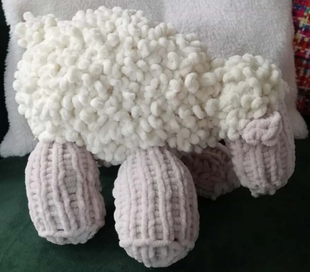 Soft handmade sheep plush with a fluffy white textured body and chunky knitted beige legs, displayed on a dark green surface.
