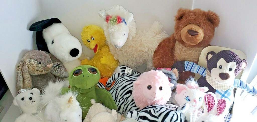A diverse group of stuffed animals, including a bear, monkey, unicorn, frog, rabbit, lamb, zebra, and cartoon characters, arranged together in a corner as a colorful plush toy collection.