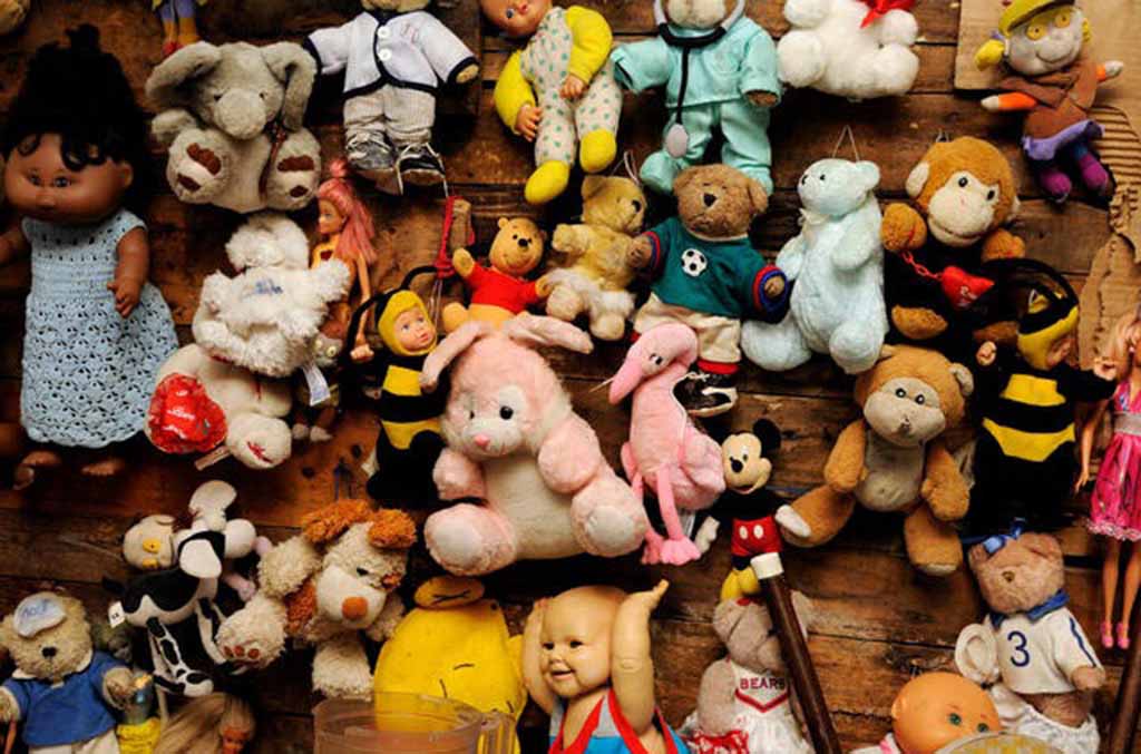 A wall filled with an eclectic mix of vintage plush toys and dolls, including teddy bears, bunnies, monkeys, flamingos, bees, and character dolls, all displayed against a rustic wooden background in a colorful, crowded arrangement.