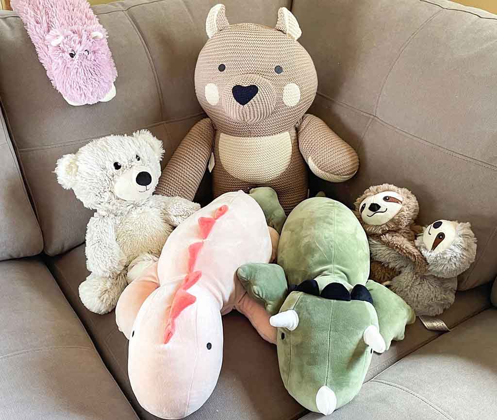 A collection of plush stuffed animals—including a knitted bear, a teddy bear, a sloth, and soft dinosaur plushies—arranged together on a couch, showcasing a mix of weighted and cuddly stuffed toys designed for comfort and relaxation.