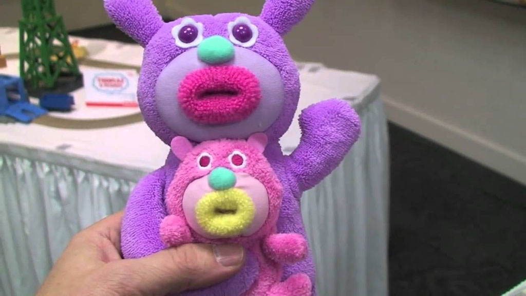 Purple plush bear holding a smaller pink baby plush bear, both with round eyes and colorful textured noses, shown close-up indoors.