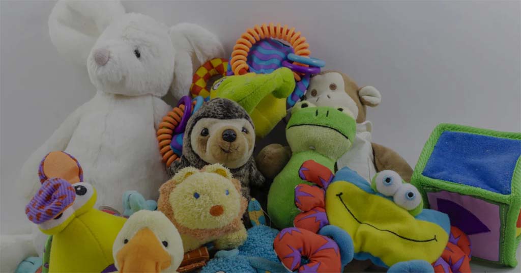 Assorted plush toys and baby soft toys piled together, including stuffed animals, rattles, and sensory plush items in bright colors