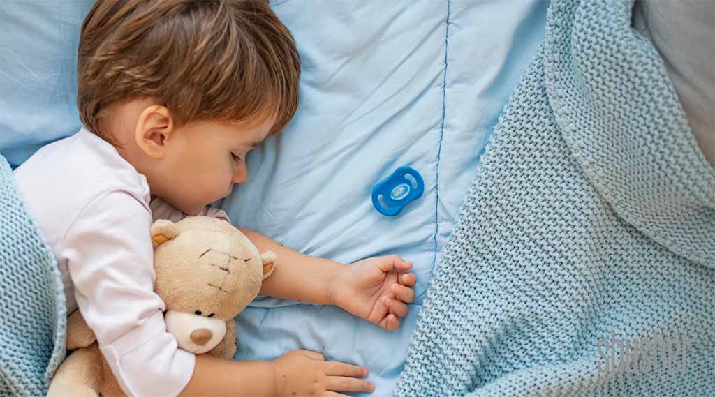 oddler sleeping peacefully on a blue bed while cuddling a soft teddy bear, with a blanket nearby.