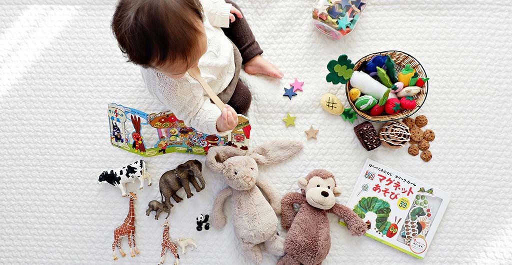Baby sitting on a soft white mat playing with plush toys, animal figures, and picture books arranged neatly around them.