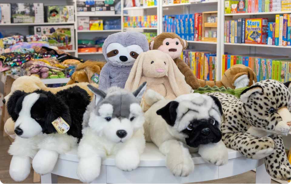 Assorted weighted stuffed animals displayed on a store table, including dogs, a sloth, a bunny, and a monkey, with colorful board games and toys on shelves in the background.