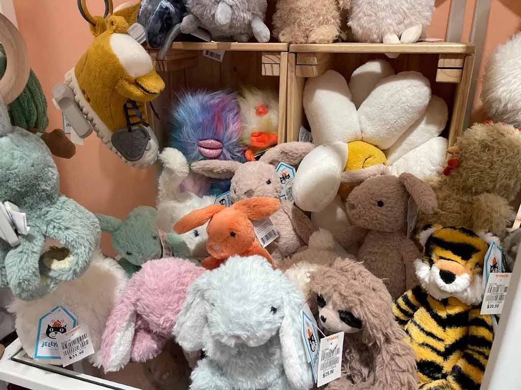 A retail display filled with assorted plush stuffed animals, including bunnies, bears, flowers, and animals in soft pastel colors, neatly arranged on wooden shelves in a toy store.