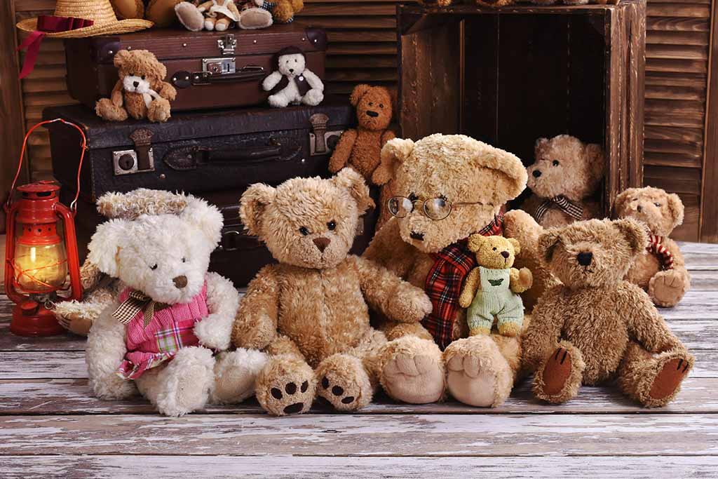 Collection of vintage-style teddy bear plush toys arranged on a rustic wooden floor, featuring classic brown and cream bears with stitched details, small accessories, and antique suitcases in the background