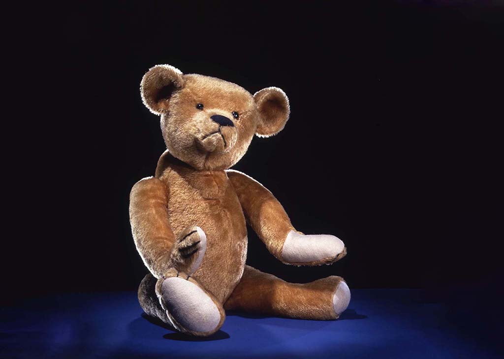 Vintage-style teddy bear sitting upright on a dark surface, with soft brown plush fabric, rounded ears, stitched facial features, and visible wear that suggests age, photographed against a black background.