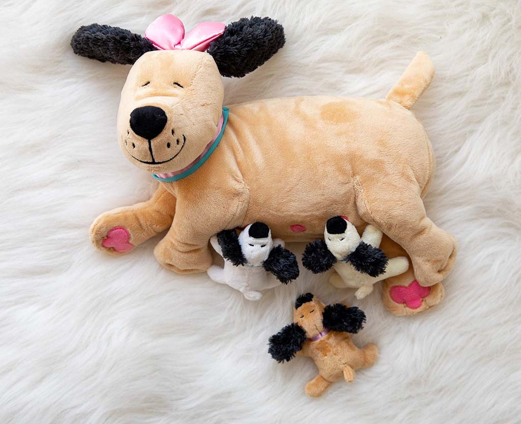 Mother dog plush toy with embroidered smile, lying on a soft white fur background, accompanied by three small puppy plush toys nestled beside her.