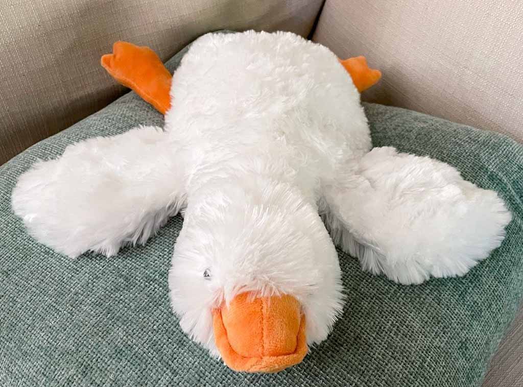 A white weighted plush duck lying flat on a green cushion, featuring a soft fluffy body, wide outstretched wings, and bright orange beak and feet, designed for calming comfort and cuddling.