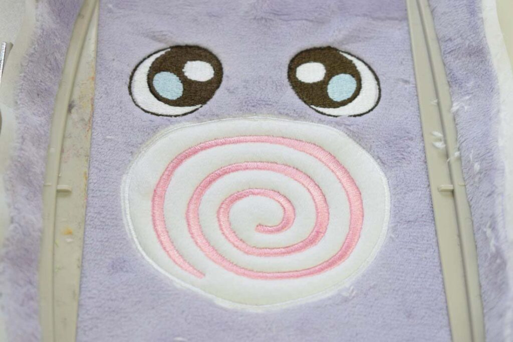 Close-up of an embroidered plush toy face inside an embroidery hoop, showing detailed stitched eyes and a spiral appliqué mouth on soft fabric.
