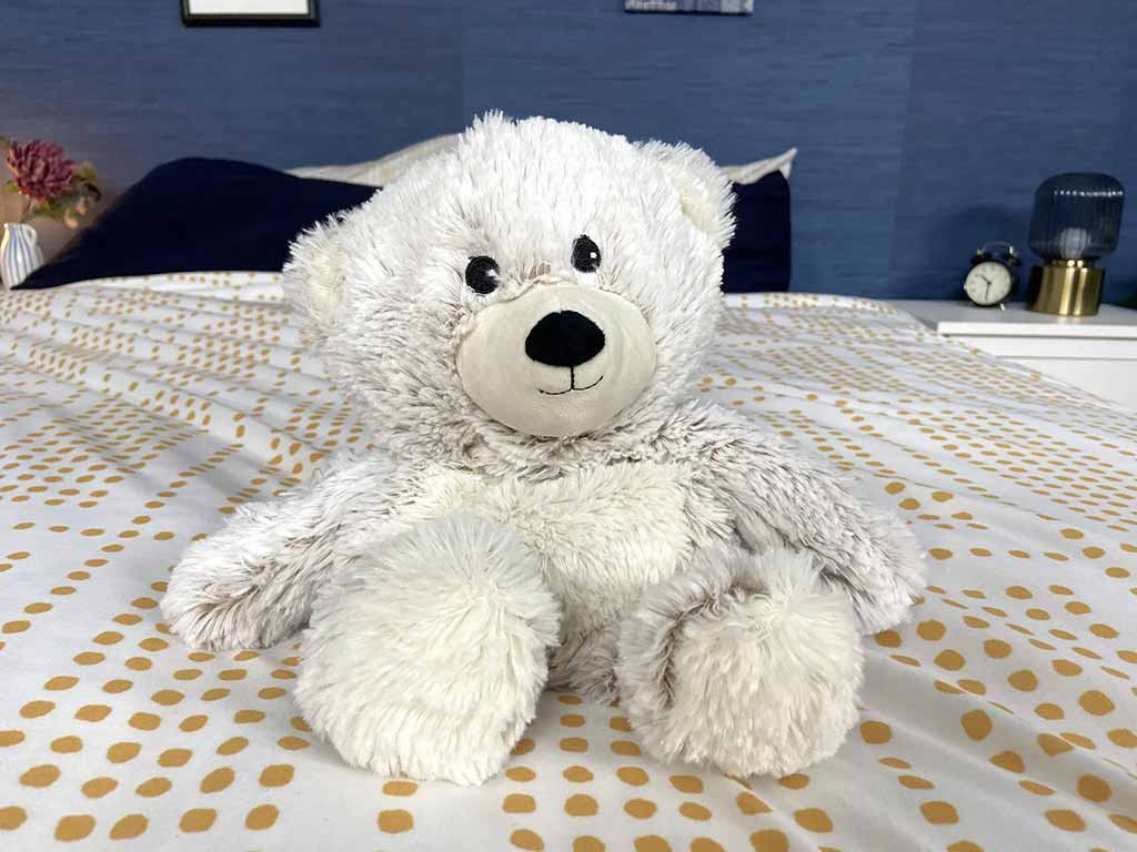 White plush teddy bear with a soft, fluffy texture sitting on a patterned bedspread, featuring a round beige snout, black stitched nose, and relaxed, cuddly posture in a cozy bedroom setting.