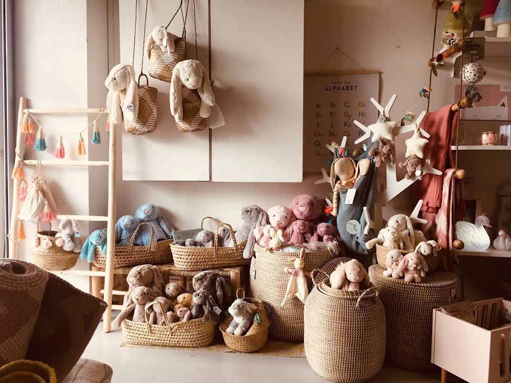 A cozy boutique-style display of plush stuffed animals arranged in woven baskets and hanging organizers, featuring soft bunny and bear plush toys in neutral pastel tones.