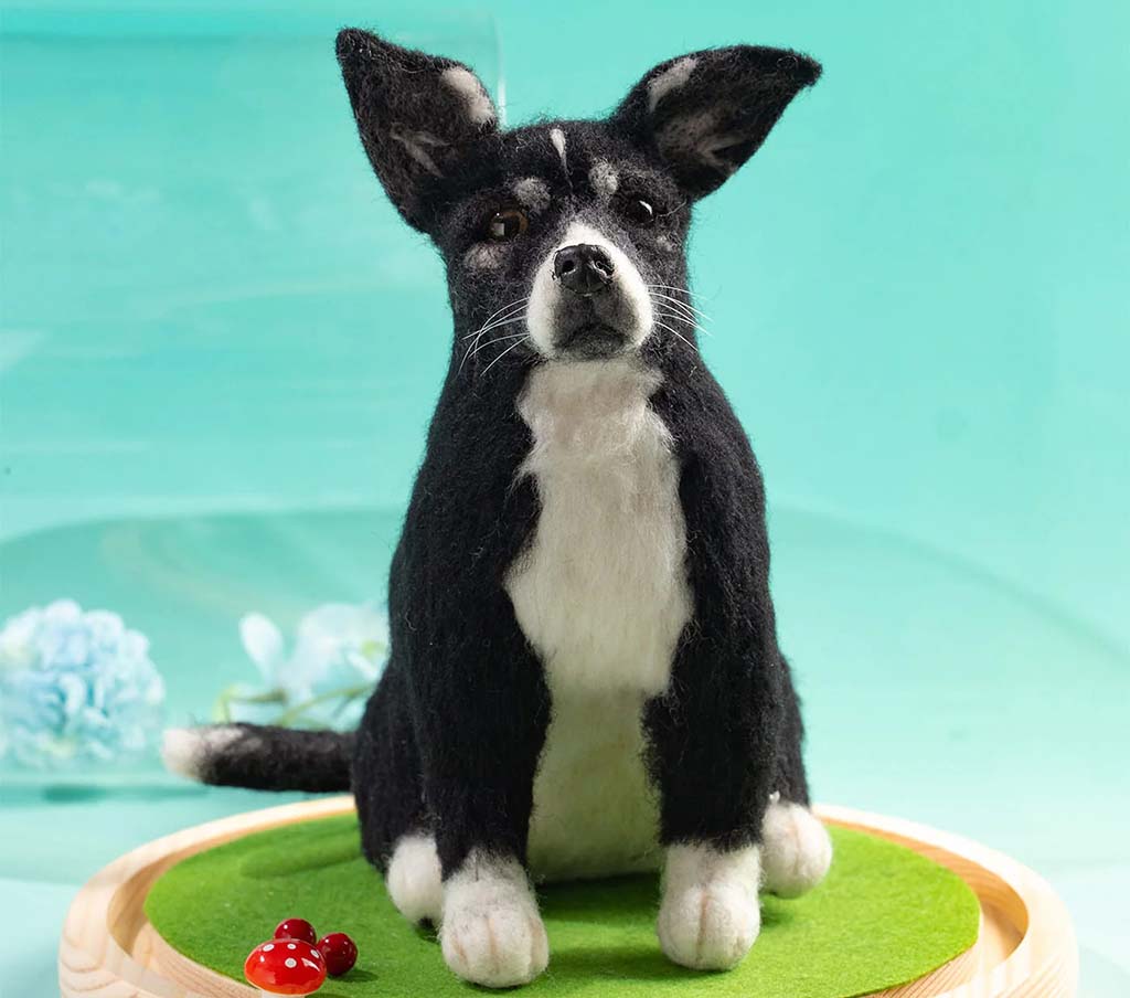 Handcrafted realistic black-and-white dog replica plush, sitting upright on a green felt display base, featuring detailed facial expression, pointed ears, and lifelike fur texture.