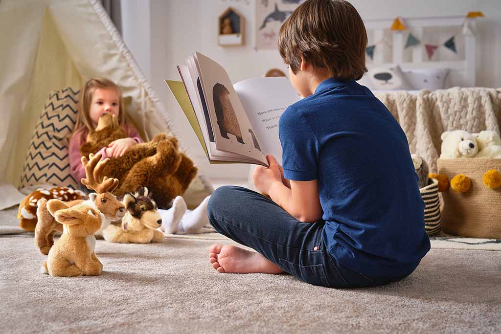 Children sitting on the floor reading a picture book together, surrounded by soft stuffed animal toys in a cozy playroom setting.