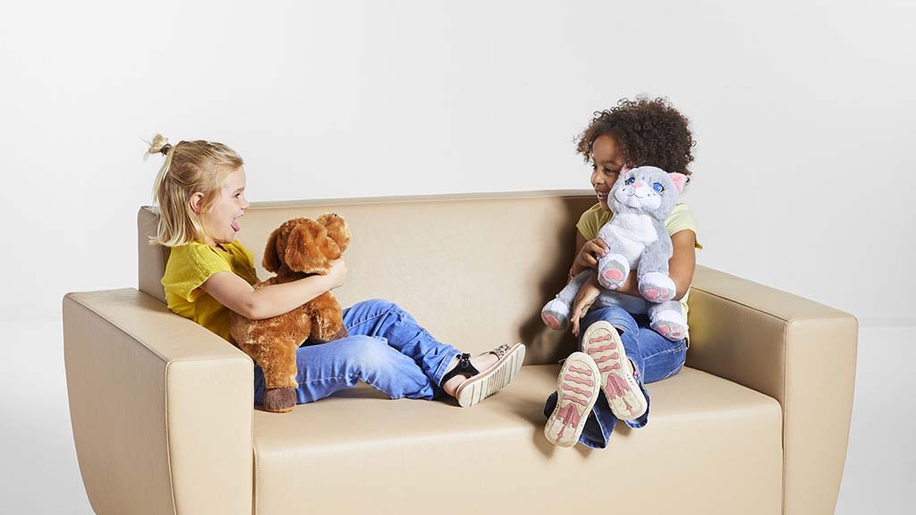 Two young children sitting on a sofa, smiling and playing together while holding soft stuffed animal toys indoors.