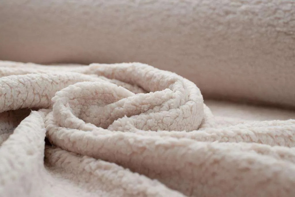Close-up of cream-colored sherpa fabric with thick, fluffy texture, softly folded to highlight warmth, loft, and cozy surface detail