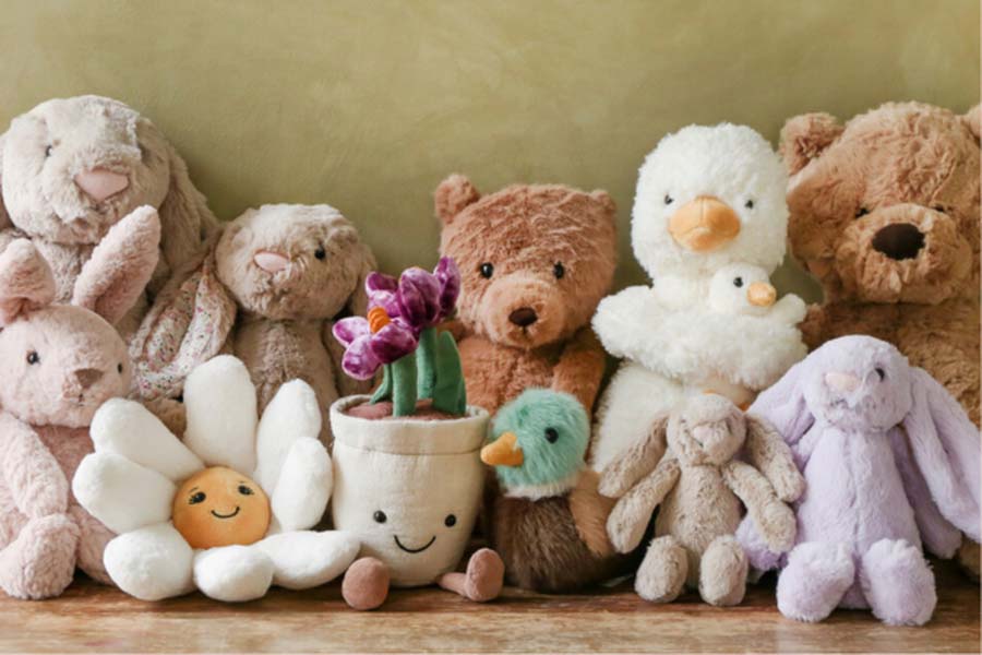 A cozy collection of Jellycat plush toys including soft bunnies, teddy bears, ducklings, and a smiling flower pot plush, arranged closely together on a wooden surface.
