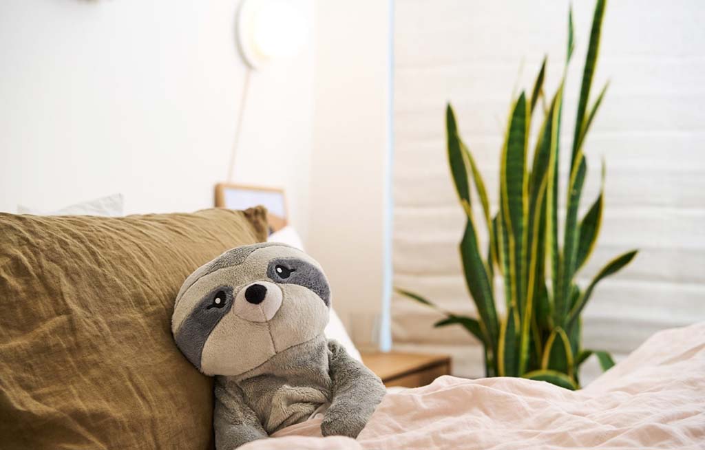 Gray sloth plush toy resting against pillows on a bed, featuring a soft weighted design, neutral bedding, and a cozy bedroom with a green indoor plant.