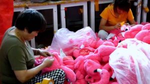 Plush toy factory workers hand-sewing and assembling bright pink stuffed animals at a production table, surrounded by partially finished plush toys and fabric materials.
